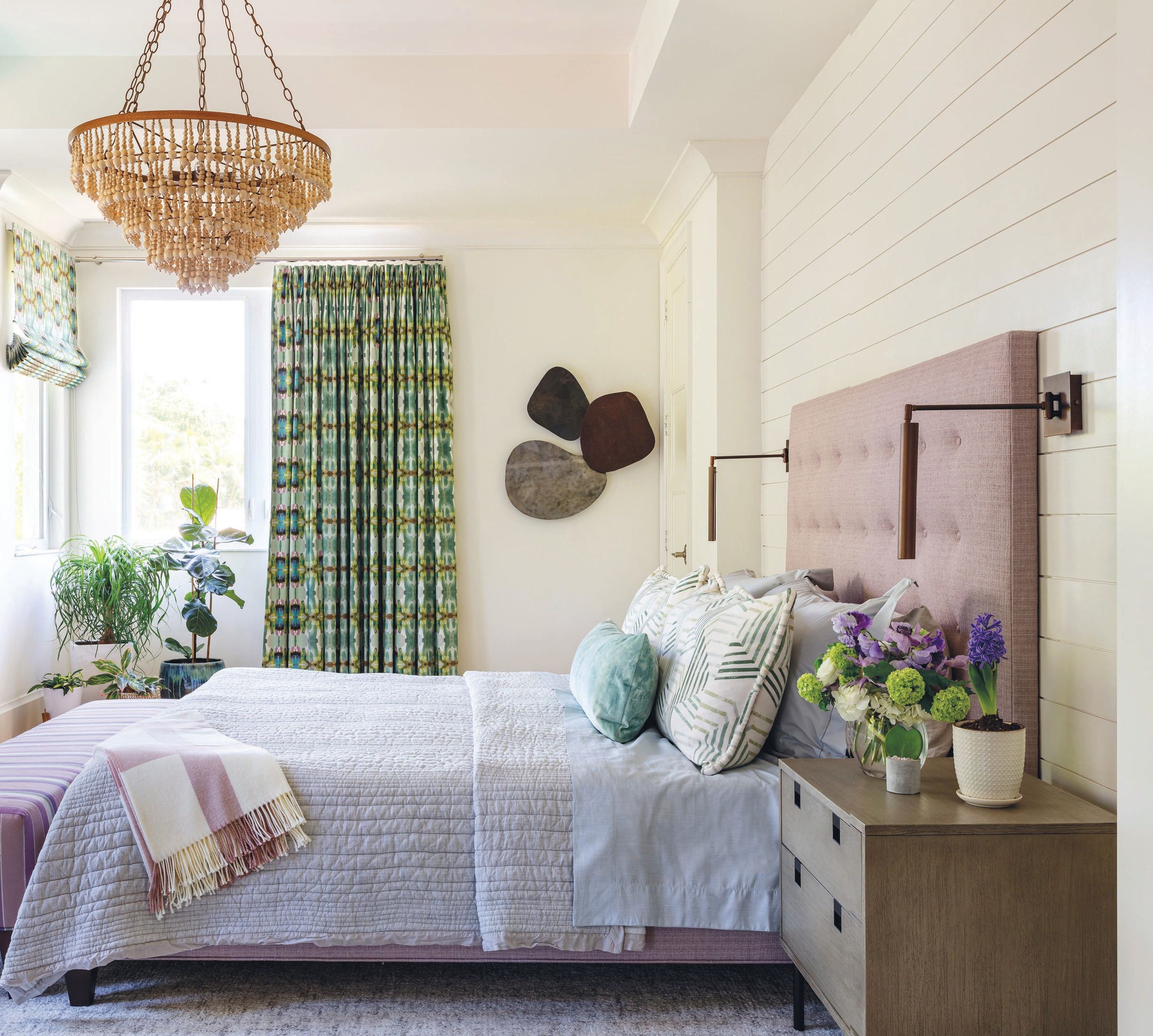 A heavy, dark ledgestone-clad wall in the primary bedroom was updated with crisp-white paneling that contrasts a bed wrapped in lilac Thibaut fabric and draperies from Laura Park Designs PHOTOGRAPHED BY NICK SARGENT